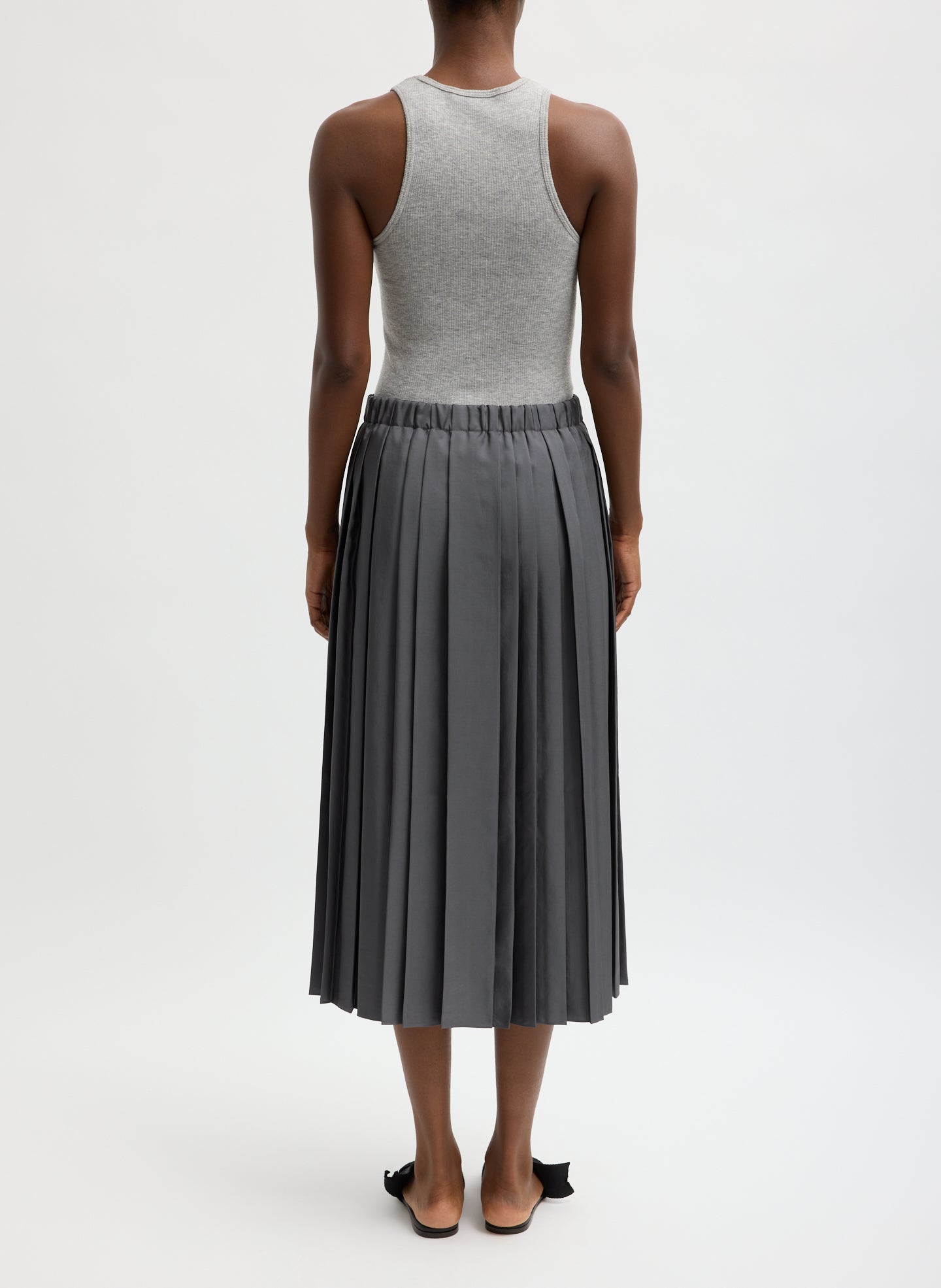 Person wearing a gray sleeveless top and pleated skirt on a white background facing the back