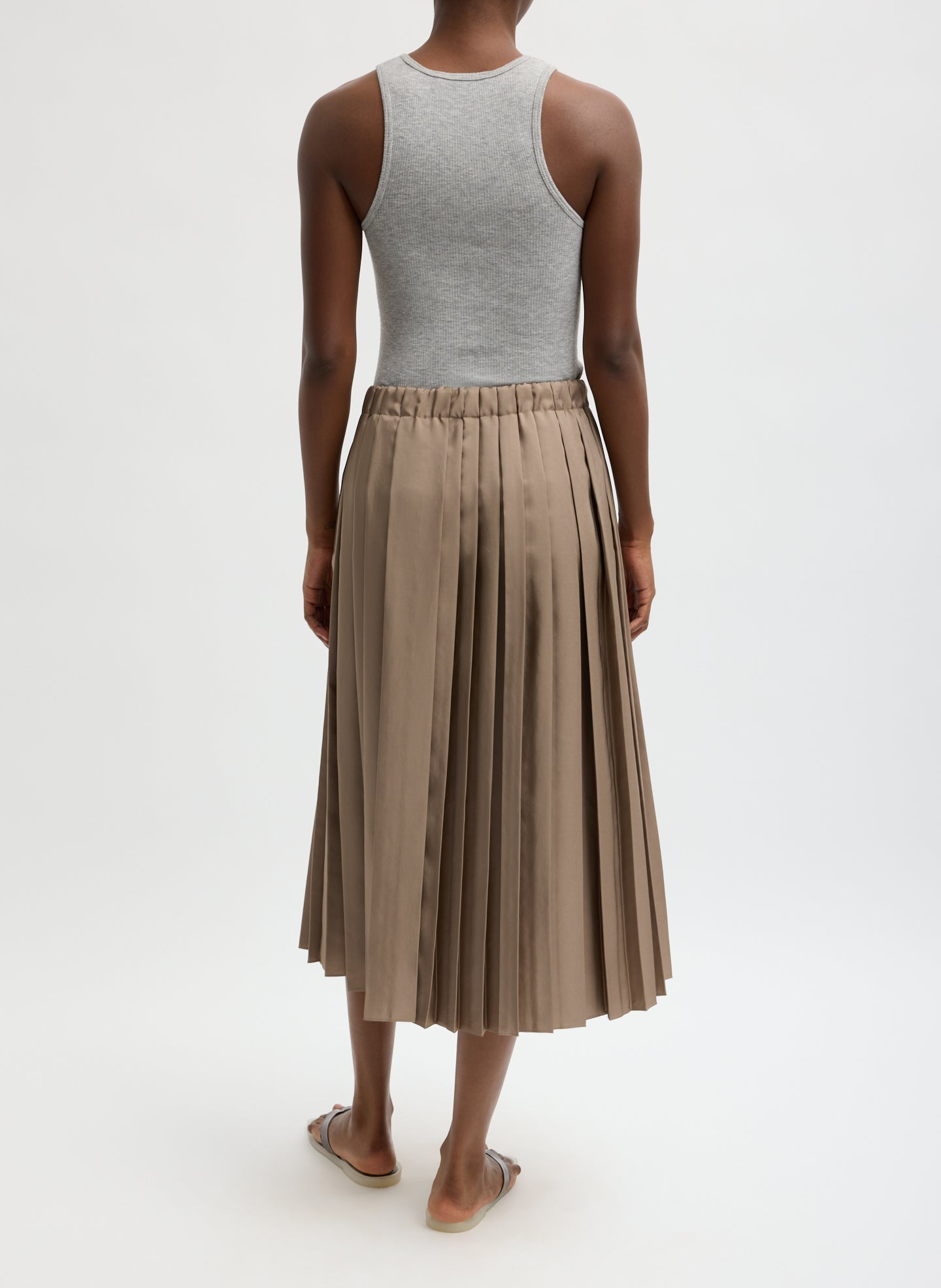 Person wearing a gray tank top and beige pleated skirt on a white background facing backwards