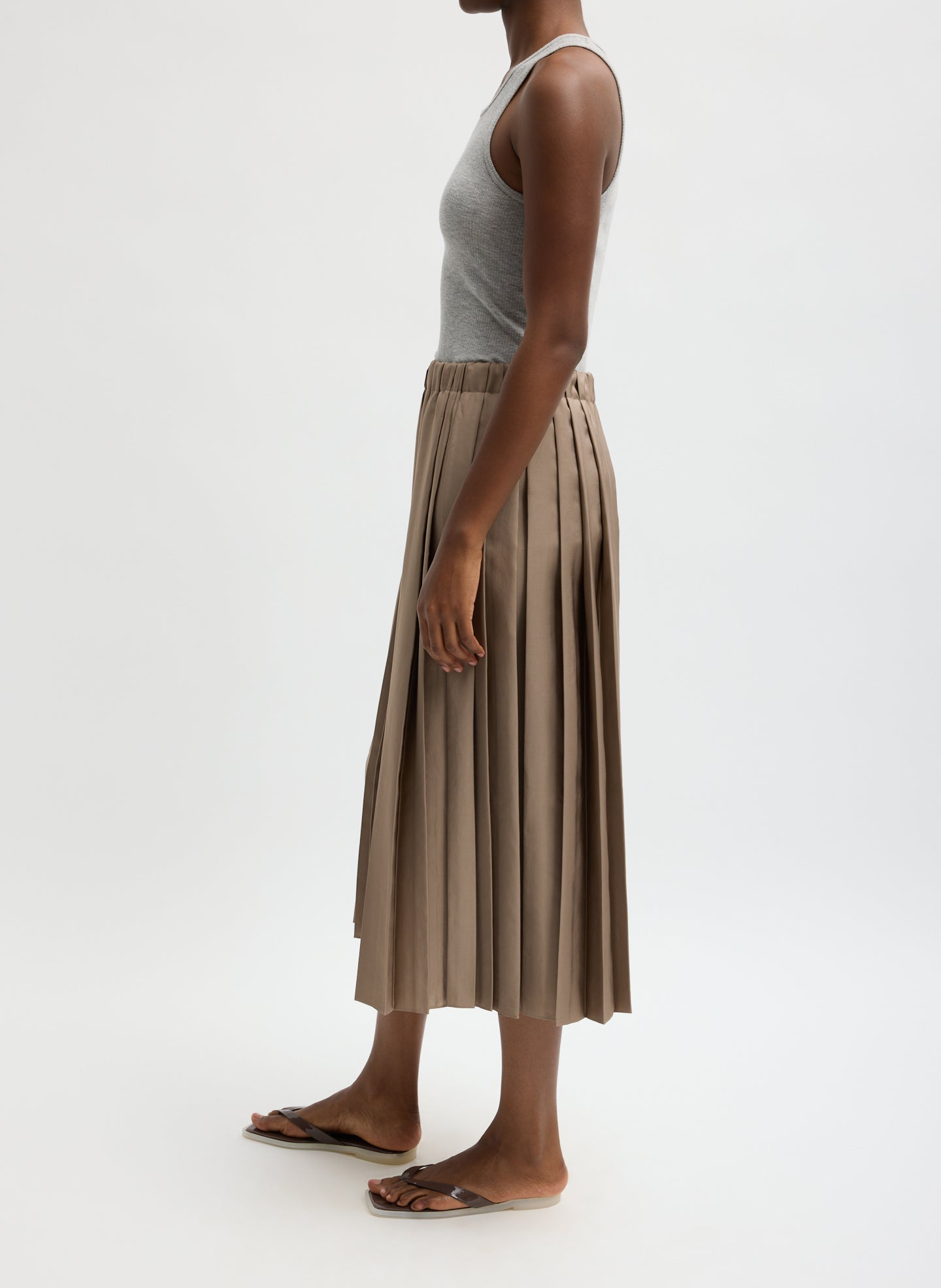Person wearing a gray tank top and brown pleated skirt on a white background turned to the side